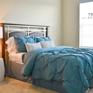 A cozy bedroom featuring a bed with blue bedding and pillows, a bedside lamp, and a window.