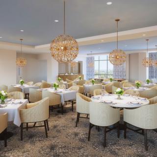 An elegant restaurant dining area with neatly arranged tables and plush chairs, illuminated by decorative chandeliers.