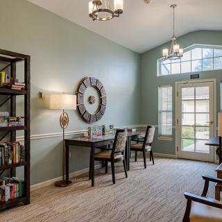 A cozy living room with bookshelves, a table with chairs, and large windows allowing natural light.