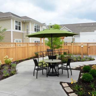 A backyard patio with outdoor furniture and a green umbrella, surrounded by a garden and enclosed by a wooden fence.