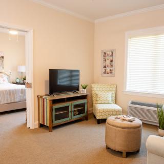 A cozy living room with a TV, chair, and window leading to a bedroom.