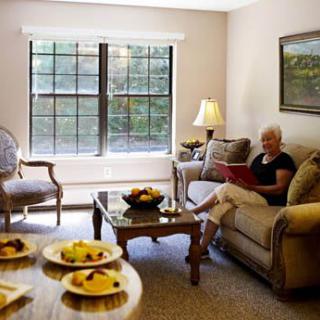 A cozy living room where a woman sits on a sofa, reading, with a window view and a table set with fruit.