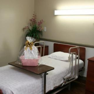 A hospital room with a neatly made bed, featuring a gift basket and flower arrangement on the table.