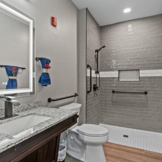 A modern bathroom with a spacious shower, sink, and toilet, featuring gray tile and accessible grab bars.