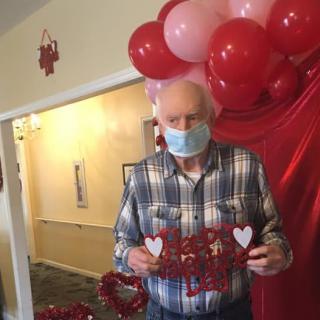 An elderly man wearing a mask holds a Valentine's Day decoration in front of festive balloons.