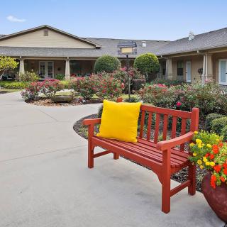 A peaceful outdoor scene with a red bench adorned with a yellow pillow, surrounded by a well-maintained garden and flowers, in front of a building.