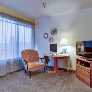 A cozy living room with a chair, desk, lamp, and television by a window with sheer curtains.