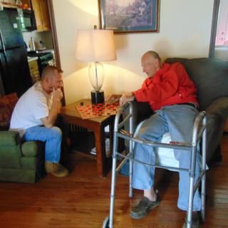 An elderly person and a younger man engage in a conversation in a cozy living room setting.