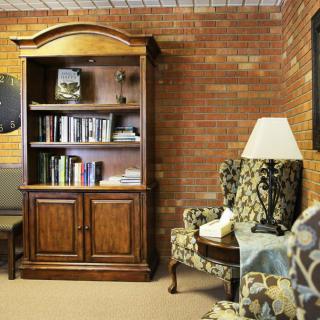 A cozy living room with a wooden bookshelf, armchair, and lamp against a brick wall.