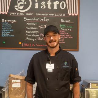 A chef standing in front of a bistro menu board, wearing a uniform and smiling.