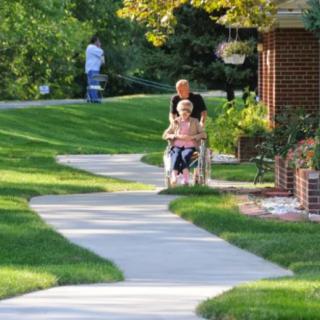 A caregiver pushing an elderly person in a wheelchair along a pathway surrounded by greenery.