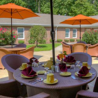 A cozy outdoor dining area featuring a round table set with colorful dishware and surrounded by wicker chairs under orange umbrellas in a garden setting.