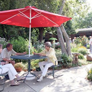 A group of people sitting at a table under a red umbrella in a lush garden setting.
