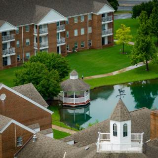 A scenic view of a residential area featuring apartments, a gazebo, and a pond surrounded by lush greenery.