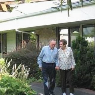 An elderly couple walks hand-in-hand in the garden outside their house.