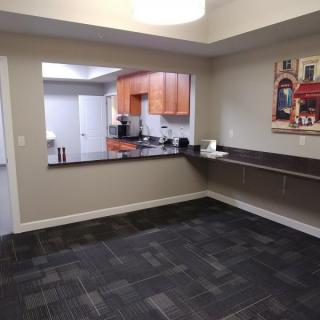 A modern office area with a view into a kitchen, featuring carpet flooring and a countertop workspace.