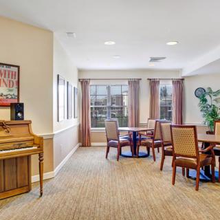 A cozy dining and lounge area featuring a piano, tables, and chairs with large windows allowing natural light.