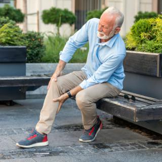 An elderly man sitting on a bench outdoors, holding his knee in discomfort.