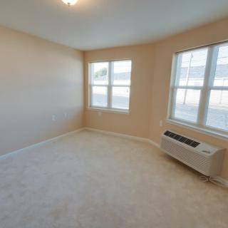 A bright, empty room with carpeted flooring and an air conditioning unit under the windows.