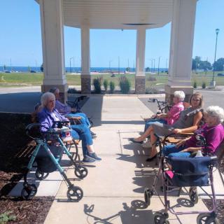 A group of seniors sitting outside on a porch, enjoying a sunny day with walkers beside them.