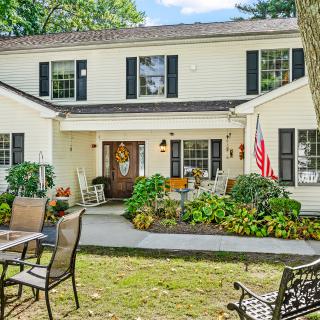 A charming white two-story house with black shutters, surrounded by a landscaped garden and outdoor patio seating.