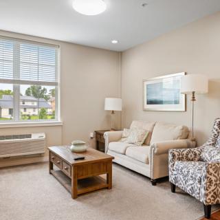 A cozy living room with a sofa, patterned armchair, and coffee table, lit by natural light from a large window.