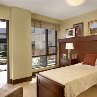 A cozy, well-lit bedroom featuring a single bed with wooden headboard, a bedside lamp, and large windows bringing in natural light.