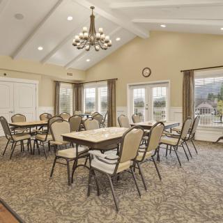 A spacious dining room with multiple tables and chairs, featuring a chandelier and large windows.