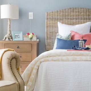 A cozy bedroom interior with a neatly arranged bed, decorative pillows, a wicker headboard, and a wooden nightstand featuring a lamp and accessories.