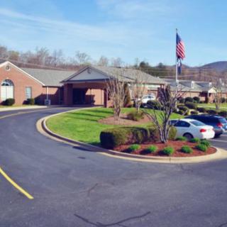 A brick building with a curved driveway and landscaped greenery, featuring several parked cars and an American flag.