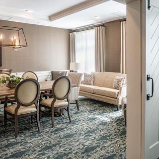A stylish dining room with a wooden table, upholstered chairs, and a comfortable sofa, complemented by an elegant carpet and modern lighting.