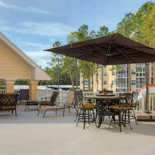 A sunny outdoor patio area with tables, chairs, and a large umbrella, surrounded by trees and buildings.