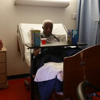 A senior patient sitting up in a hospital bed, surrounded by care items.
