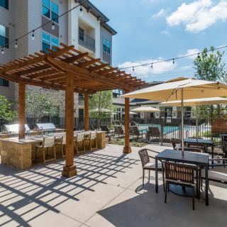 An outdoor patio area with a pergola, tables, chairs, and umbrellas on a sunny day.