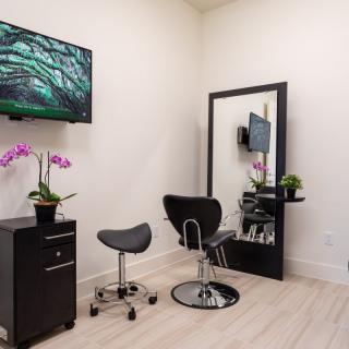 A modern salon interior featuring a styling chair, mirror, decorative plants, and a television on the wall.