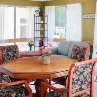 A cozy living room with a wooden dining table surrounded by chairs featuring floral upholstery, accented by a vase of flowers and sunlight streaming through the windows.
