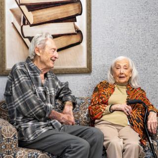 An elderly couple sitting together, engaged in conversation beside a painting of books.