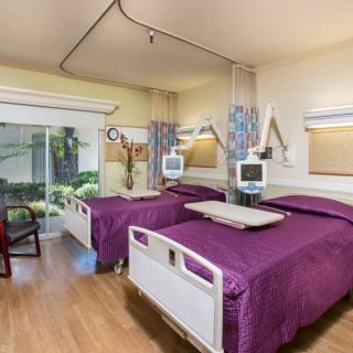 A modern hospital room featuring twin beds with medical equipment and a view of greenery outside.