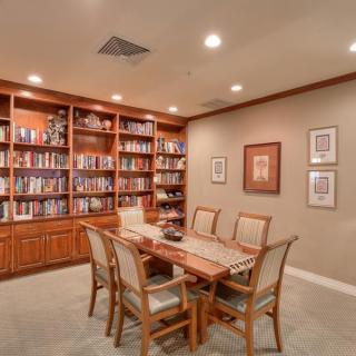 A cozy library room with wooden bookshelves filled with books, a large table, and chairs arranged neatly on a carpeted floor.