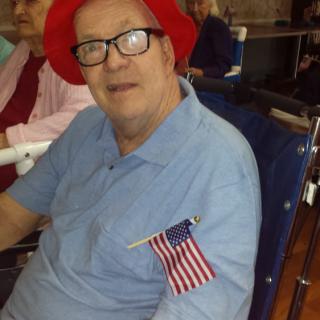 A man wearing a red hat and glasses is seated in a wheelchair, with an American flag pinned to his blue shirt.