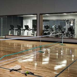 An indoor pickleball court with paddles on the floor, adjacent to a gym with exercise equipment.