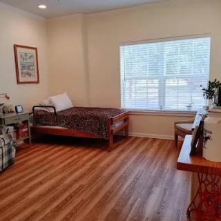 A cozy and simple room with a bed, a small table, and chairs, featuring natural light from a window.