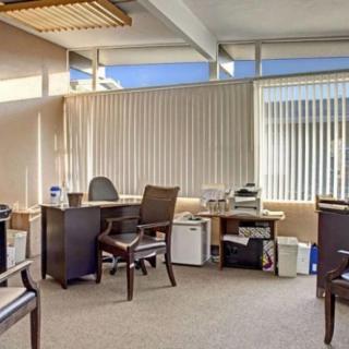 A well-lit office space with desks and chairs, featuring large windows with blinds.