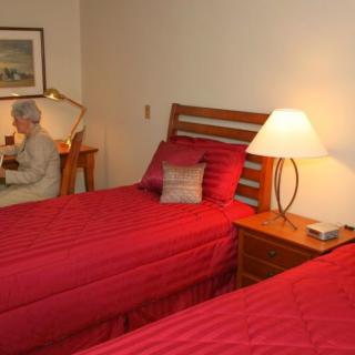 A cozy bedroom with two twin beds covered in red bedding, a lamp on the nightstand, and an elderly person sitting at a desk.