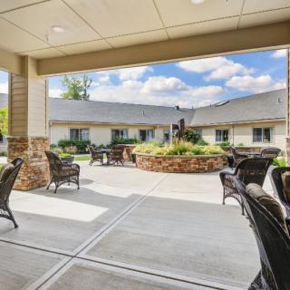 A covered outdoor patio with seating, overlooking a landscaped courtyard and building under a blue sky.