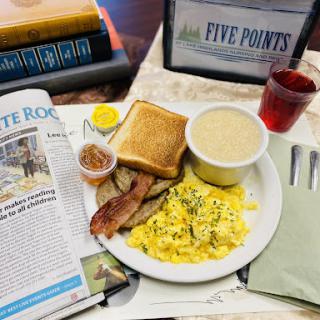 A hearty breakfast spread featuring scrambled eggs, toast, bacon, grits, and a side of newspaper reading.