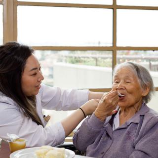 A caregiver assists an elderly woman by feeding her in a comfortable setting.