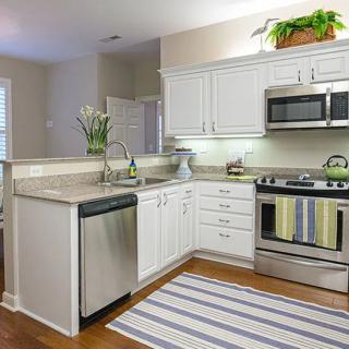 A modern kitchen with white cabinets and stainless steel appliances, featuring a dishwasher, oven, microwave, wooden flooring, and a striped rug.