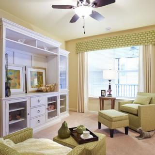 A cozy living room with light green armchairs and a well-organized bookshelf, illuminated by natural light from a large window.