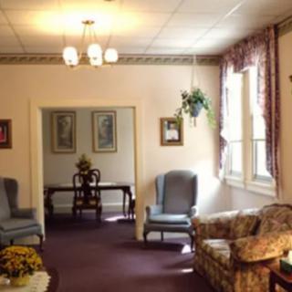 A cozy living room with vintage-style furniture, including armchairs and a floral sofa, complemented by elegant decor and natural lighting from large windows.